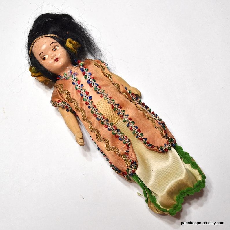 Puede incluir: Una mu&ntilde;eca vintage con cabello negro y un vestido color melocot&oacute;n adornado con detalles dorados y coloridos. El vestido de la mu&ntilde;eca tiene una falda color crema con un ribete verde. La mu&ntilde;eca est&aacute; sobre una superficie blanca.