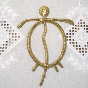 Peut inclure: Une décoration murale en métal doré représentant une figure humaine stylisée et abstraite avec un torse circulaire, une petite tête ronde, des bras étendus et trois jambes pointant vers le bas, le tout avec une finition martelée texturée.
