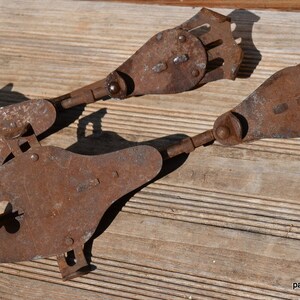 May include: A pair of antique metal ice skates featuring a heavily rusted, weathered brown finish. The skates have a distinct elongated shape with a rounded front, a central cutout, and a mechanical heel adjustment mechanism with a metal plate.