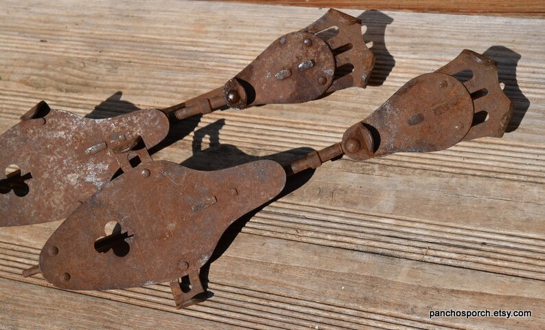 May include: A pair of antique metal ice skates featuring a heavily rusted, weathered brown finish. The skates have a distinct elongated shape with a rounded front, a central cutout, and a mechanical heel adjustment mechanism with a metal plate.