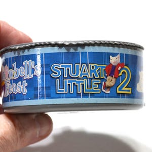 May include: A cylindrical metal can with a blue plaid background featuring the text Stuart Little 2 and a graphic of a white mouse character wearing a red jacket. The label also includes the text Furr-well's Feast in a stylized blue font.