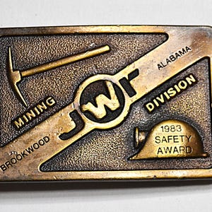 May include: Bronze-coloured rectangular belt buckle with a textured surface. It features the text "BROOKWOOD MINING DIVISION ALABAMA" and "1983 SAFETY AWARD". Includes a pickaxe, a hard hat, and the letters "JWF".
