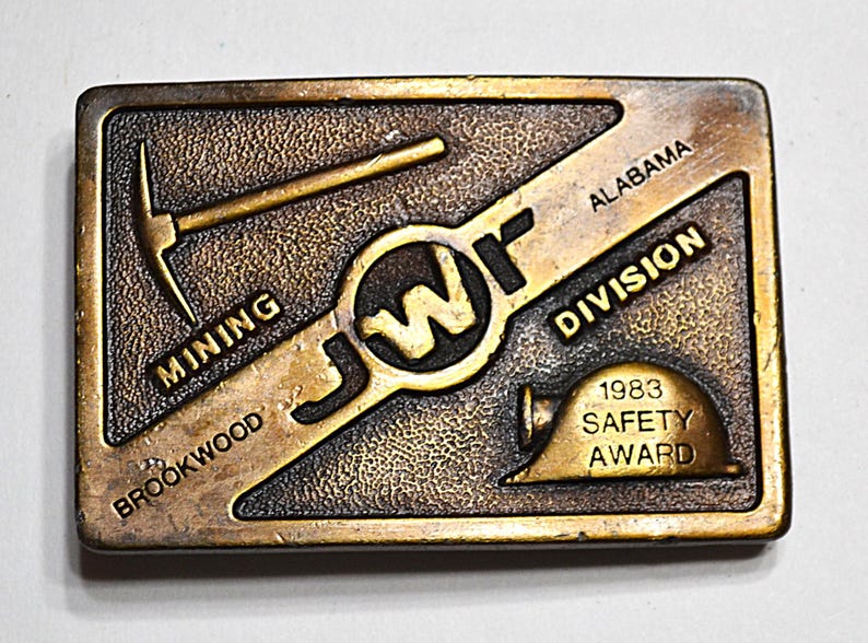 May include: Bronze-coloured rectangular belt buckle with a textured surface. It features the text "BROOKWOOD MINING DIVISION ALABAMA" and "1983 SAFETY AWARD". Includes a pickaxe, a hard hat, and the letters "JWF".