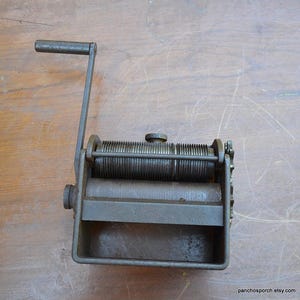 May include: An antique cast iron pasta maker with a hand crank. The machine is a dark, aged metal with a rectangular base and a series of rollers. The crank handle is attached to the side, and the machine sits on a wooden surface.