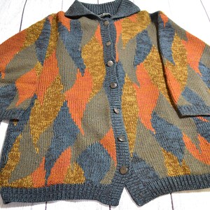 May include: A long-sleeve knit cardigan featuring a bold, abstract diamond pattern in shades of burnt orange, mustard yellow, and slate blue. The garment includes a dark grey ribbed collar, cuffs, and hem, with a front button closure down the center.