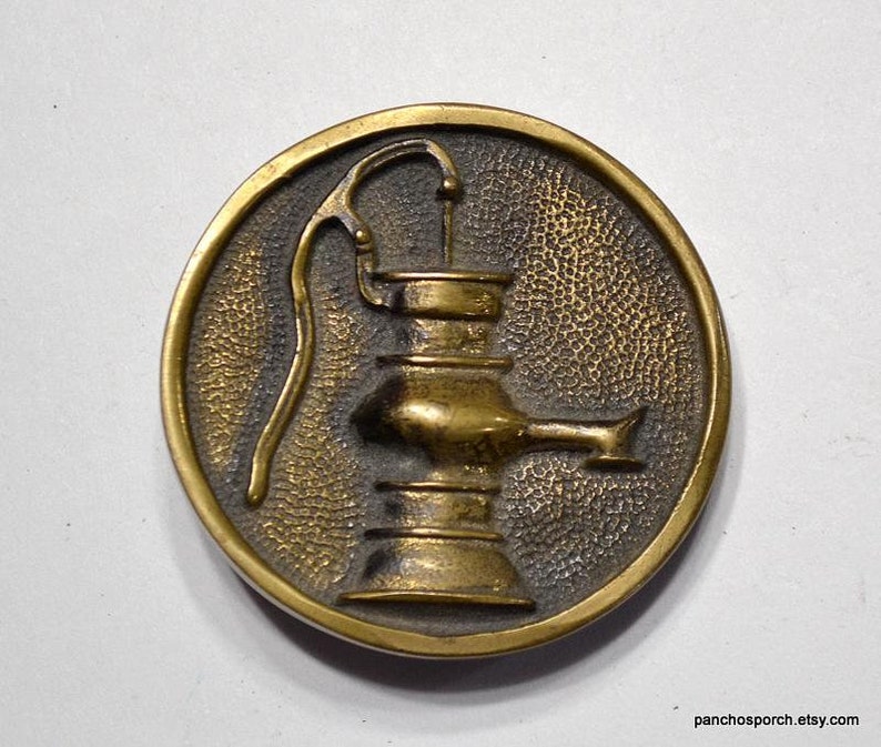 K&ouml;nnte beinhalten: Runder Metallknopf im antiken Stil mit einem erhabenen Design einer Wasserpumpe. Der Knopf hat einen strukturierten Hintergrund und eine goldfarbene Oberfl&auml;che mit einem dunkleren, gealterten Aussehen. Die Pumpe ist mit Griff und Ausguss detailliert.