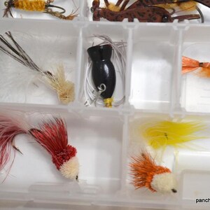 May include: A clear plastic tackle box filled with various colorful fishing lures. The lures include a black lure, red and white lures, and yellow and orange lures. The lures are designed with feathers and other materials.
