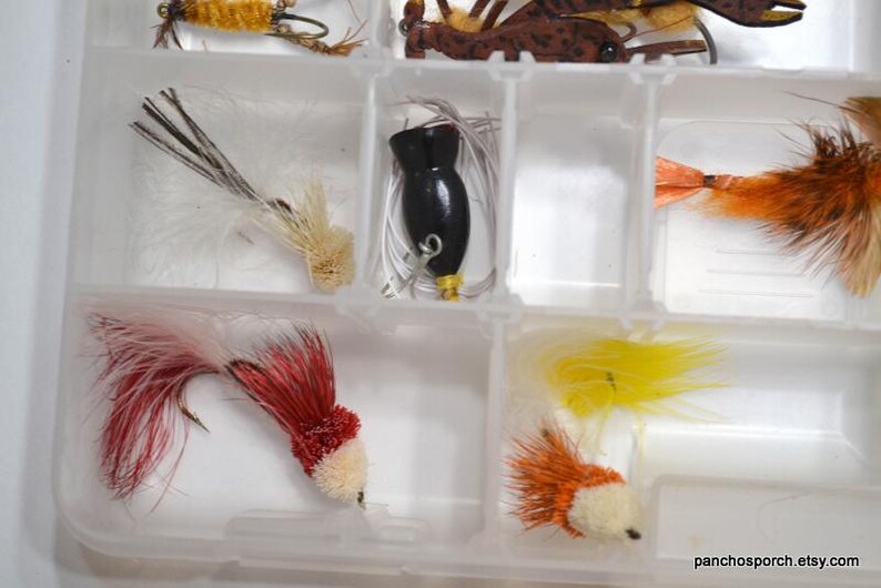 May include: A clear plastic tackle box filled with various colorful fishing lures. The lures include a black lure, red and white lures, and yellow and orange lures. The lures are designed with feathers and other materials.
