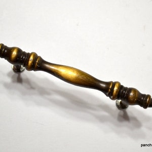 May include: A metal drawer pull handle featuring a dark antique brass finish with a central spindle shape and decorative rounded ends. The hardware has a smooth, polished texture with darker recessed areas and lighter highlights on the curves.