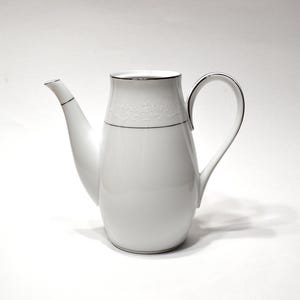 May include: A white porcelain coffee pot featuring a smooth, rounded body, a curved handle, and a tapered spout. The upper section is decorated with a subtle, embossed floral pattern and a thin silver rim along the top edge and spout.
