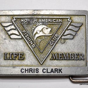 May include: Silver belt buckle with a North American Fishing Club logo featuring a fish and the words "Life Member" and "Chris Clark".