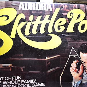 May include: A rectangular cardboard game box for Aurora Skittle Pool featuring large yellow text on a black background. The box displays a photograph of a person using a cue to aim a black ball through a triangular frame on a pool table surface.