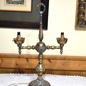Puede incluir: Una l&aacute;mpara candelabro de metal de dos luces con un acabado en oro y plata desgastado. La l&aacute;mpara presenta un poste central vertical con un asa de anillo en la parte superior, dos casquillos estilo vela con arandelas en forma de hoja.