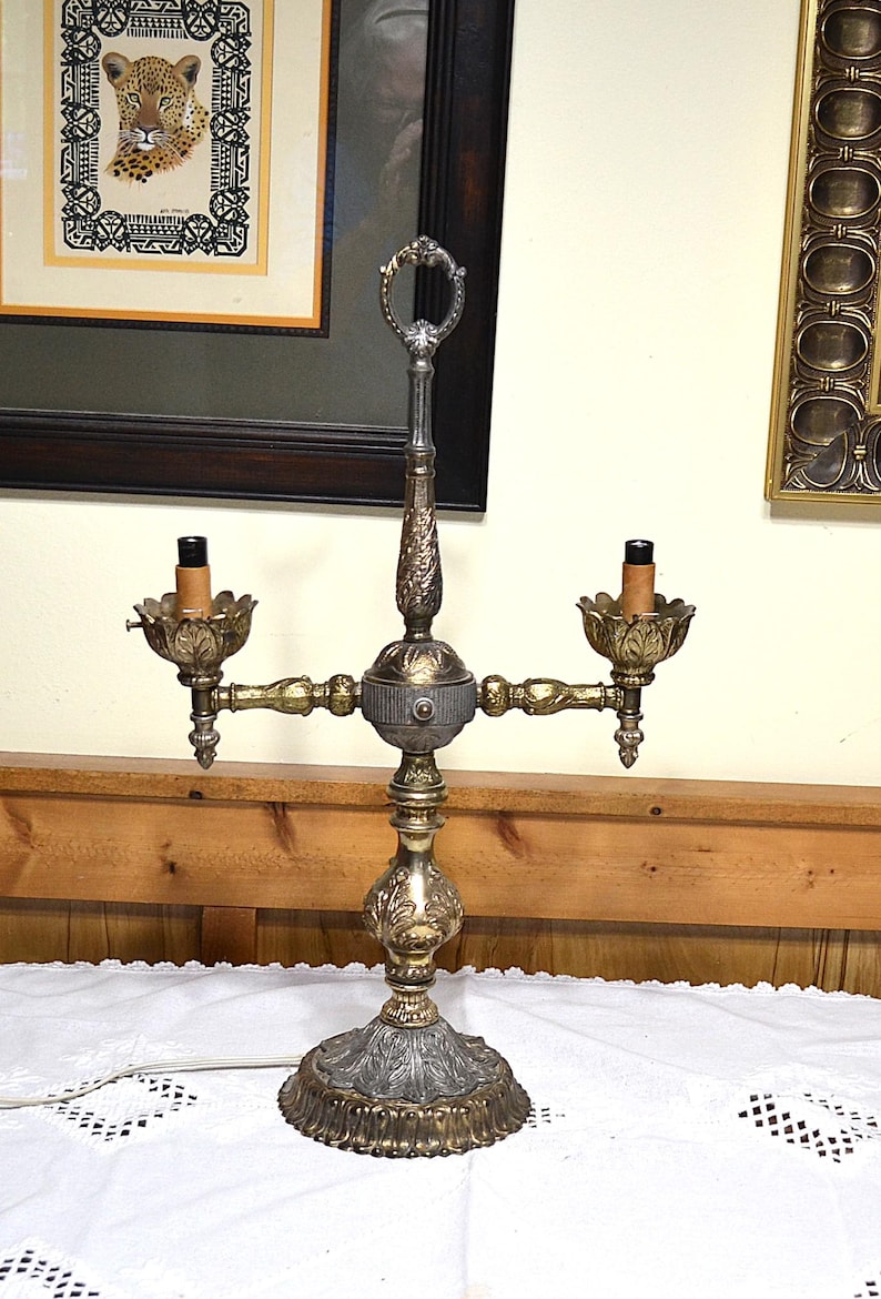 Puede incluir: Una l&aacute;mpara candelabro de metal de dos luces con un acabado en oro y plata desgastado. La l&aacute;mpara presenta un poste central vertical con un asa de anillo en la parte superior, dos casquillos estilo vela con arandelas en forma de hoja.