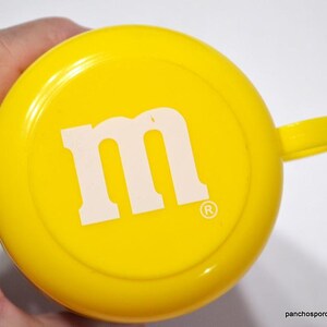May include: Bright yellow cup with a handle, featuring a large white "M" logo in the center. The cup has a rounded shape and a small handle. The design is simple and eye-catching.