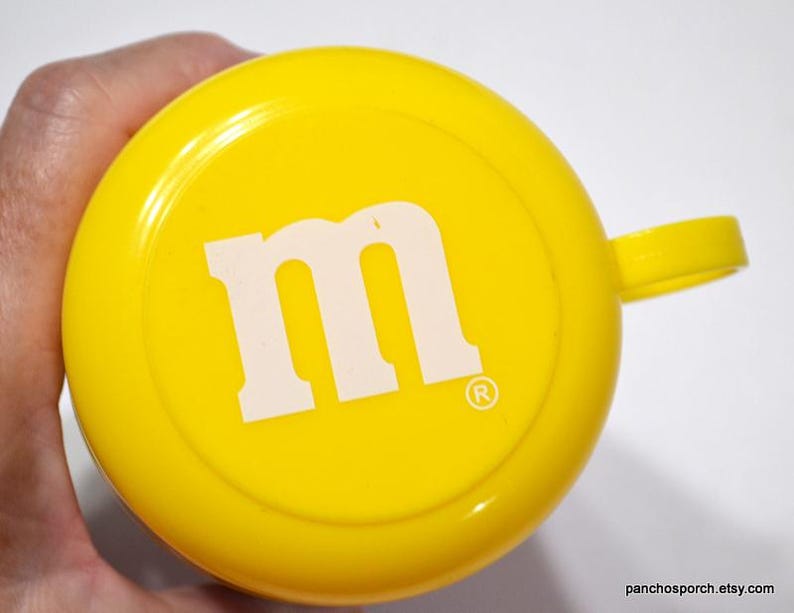 May include: Bright yellow cup with a handle, featuring a large white "M" logo in the center. The cup has a rounded shape and a small handle. The design is simple and eye-catching.