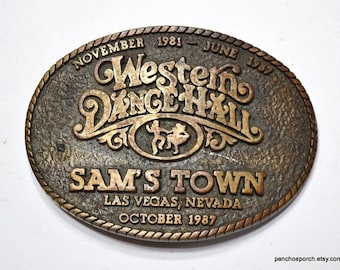 Puede incluir: Hebilla de cinturón ovalada de color bronce con las palabras "Western Dance Hall" y "Sam's Town Las Vegas, Nevada October 1987" en relieve. La hebilla tiene un borde en forma de cuerda y las fechas de noviembre de 1981 a junio de 1982.