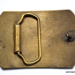 May include: A vintage brass belt buckle with a rectangular shape and rounded edges. The buckle features a loop and a pin for securing the belt. The surface has a textured pattern and some signs of wear.