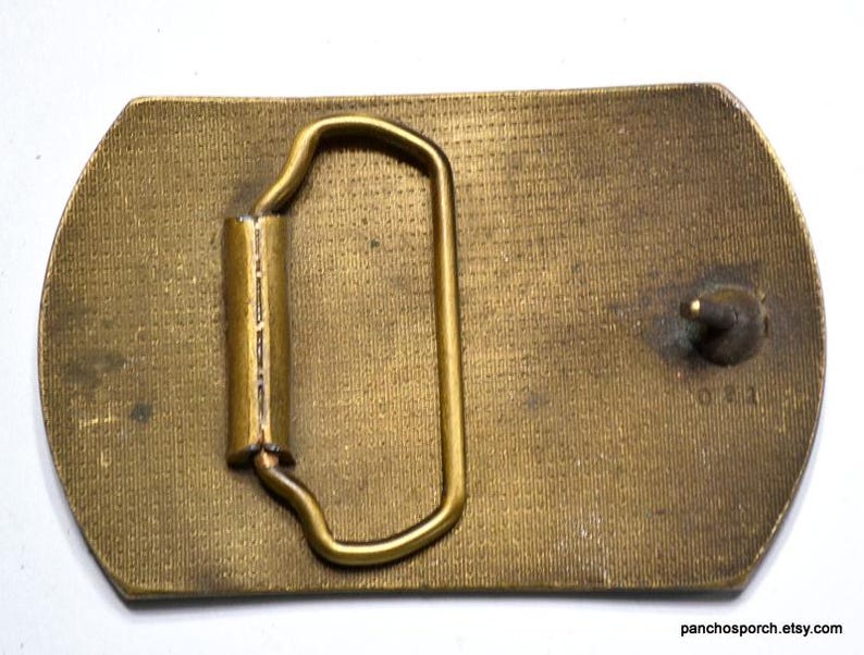 May include: A vintage brass belt buckle with a rectangular shape and rounded edges. The buckle features a loop and a pin for securing the belt. The surface has a textured pattern and some signs of wear.