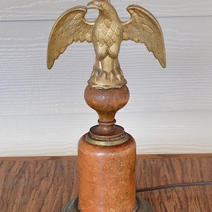 May include: A vintage table lamp featuring a gold-colored eagle with outstretched wings atop a wooden base. The lamp has a cylindrical wooden body and a brass-colored base and accents. The lamp is designed for indoor use.