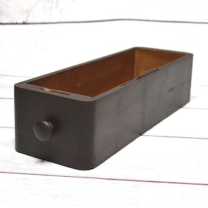 May include: A rectangular wooden drawer with a dark brown exterior finish and a lighter natural wood interior. The front panel features a single small, round, dark brown wooden knob centered on the face, providing a simple handle for opening.