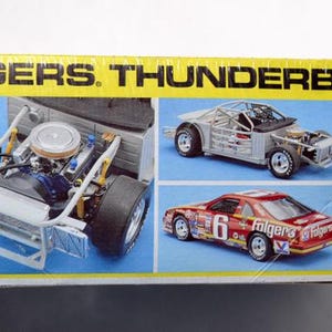 Puede incluir: Caja de modelo de un coche de carreras Folgers Thunderbird. La caja presenta im&aacute;genes del motor, el chasis y el modelo terminado del coche. La caja es amarilla con el texto "FOLGERS. THUNDERBIRD" y el logotipo de Monogram.