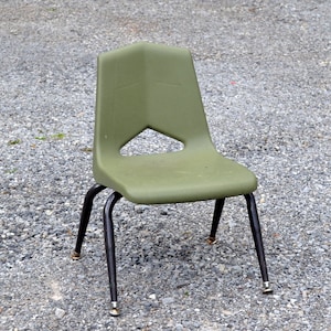 May include: A green plastic chair with black metal legs and gold-colored tips. The chair is sitting on a gravel surface.
