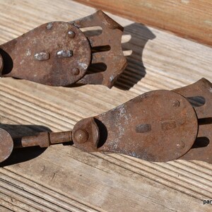 May include: A pair of antique metal ice skates featuring a rusted brown finish, a flat blade-like base with a decorative cutout design, and a hinged mechanism connecting to a long, thin metal rod, showing signs of age and oxidation throughout.