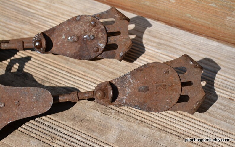 May include: A pair of antique metal ice skates featuring a rusted brown finish, a flat blade-like base with a decorative cutout design, and a hinged mechanism connecting to a long, thin metal rod, showing signs of age and oxidation throughout.