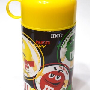 May include: A yellow and black M&M's themed container with a yellow lid and handle. The container features various M&M's characters and the text "RED" and "m&m's". The container is cylindrical.
