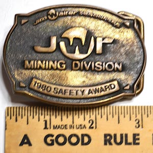 May include: Vintage brass-toned belt buckle with the text "JWP MINING DIVISION 1980 SAFETY AWARD." The buckle is rectangular with rounded edges and a detailed design. A wooden ruler with inch markings is below the buckle.