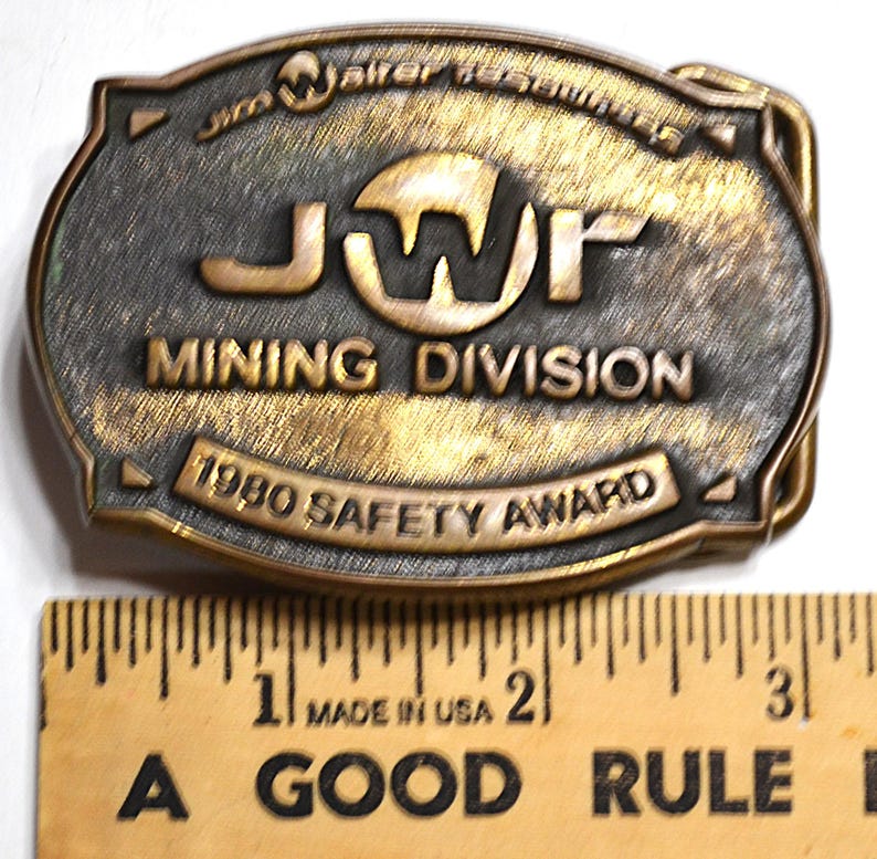 May include: Vintage brass-toned belt buckle with the text "JWP MINING DIVISION 1980 SAFETY AWARD." The buckle is rectangular with rounded edges and a detailed design. A wooden ruler with inch markings is below the buckle.