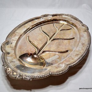 May include: An oval, silver-plated serving platter with a decorative tree-shaped carving and a built-in gravy boat. The platter has a scalloped edge with a rope-like border. The surface has a slightly tarnished appearance, adding to its vintage charm.