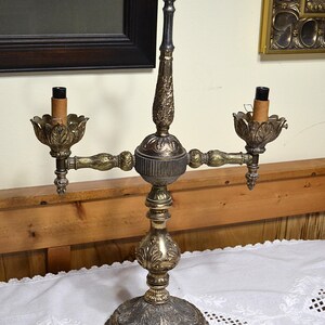 Puede incluir: Una l&aacute;mpara candelabro de metal con dos luces, que presenta una columna central texturizada y ornamentada con un asa de anillo decorativa en la parte superior. La base y los portavelas muestran intrincados patrones de hojas en acabado dorado y plateado.