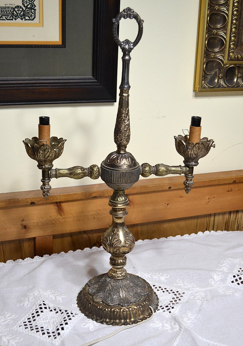 Puede incluir: Una l&aacute;mpara candelabro de metal con dos luces, que presenta una columna central texturizada y ornamentada con un asa de anillo decorativa en la parte superior. La base y los portavelas muestran intrincados patrones de hojas en acabado dorado y plateado.