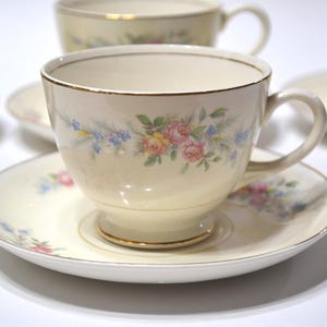 May include: Cream-colored teacups and saucers with floral designs. The cups and saucers have a gold trim. The floral design includes pink, yellow, and blue flowers. The teacups have a handle.