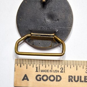 K&ouml;nnte beinhalten: Vintage Metallschnalle mit kreisf&ouml;rmigem Design und messingfarbenem Rahmen. Die Schnalle ist dunkelgrau mit strukturierter Oberfl&auml;che. Die W&ouml;rter "MADE IN USA" und "A GOOD RULE" sind auf einem Holzlineal aufgedruckt.