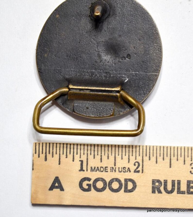 K&ouml;nnte beinhalten: Vintage Metallschnalle mit kreisf&ouml;rmigem Design und messingfarbenem Rahmen. Die Schnalle ist dunkelgrau mit strukturierter Oberfl&auml;che. Die W&ouml;rter "MADE IN USA" und "A GOOD RULE" sind auf einem Holzlineal aufgedruckt.