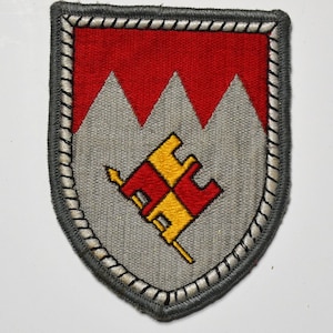 May include: A shield-shaped patch with a red upper section featuring three gray mountain peaks. The lower section is gray with a red and yellow emblem. The patch is bordered with a gray and white stitched design.