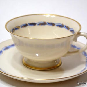 May include: Cream-colored teacup and saucer set with gold trim and blue floral accents. The teacup has a rounded shape and a small handle. The saucer is round and matches the teacup's design. A classic tea set.