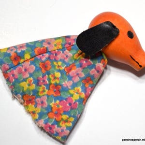 May include: A wooden puppet with a floral fabric dress and a bright orange head with a black leather ear. The puppet has a small black eye and a carved mouth.