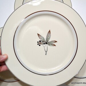 May include: Set of four white ceramic dinner plates with a gold rim and a stylized leaf design in shades of green and brown.