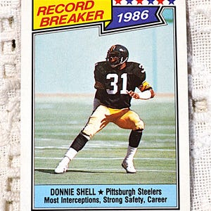 May include: A 1986 Record Breaker sports trading card featuring football player Donnie Shell of the Pittsburgh Steelers. The card shows the athlete in a black and gold uniform with the number 31, noting his career record for most interceptions.
