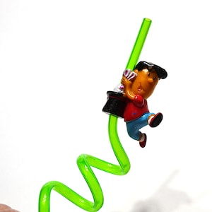 May include: A green, flexible straw with a cartoon figure of a magician holding a top hat. The magician is wearing a red shirt, blue pants, and a black hat. The straw is curved and has a bright green color.