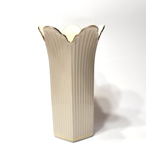 May include: A cream-colored porcelain vase with a vertical ribbed texture and a scalloped rim. The top edge and the base are accented with a thin gold trim. The vase has a flared opening and a square-shaped base, standing upright on a surface.