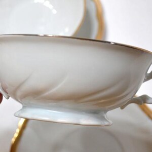 May include: White porcelain teacup with gold trim and a decorative handle. The cup has a delicate, embossed design on the side. A matching saucer is partially visible in the background. This elegant teacup is perfect for serving tea.
