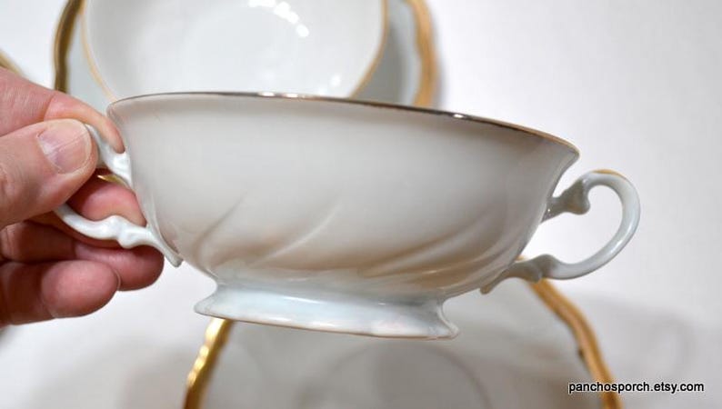 May include: White porcelain teacup with gold trim and a decorative handle. The cup has a delicate, embossed design on the side. A matching saucer is partially visible in the background. This elegant teacup is perfect for serving tea.