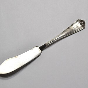 May include: A silver butter knife with a curved blade and a decorative handle. The handle features a floral design and an initial. The knife is set against a gray background.