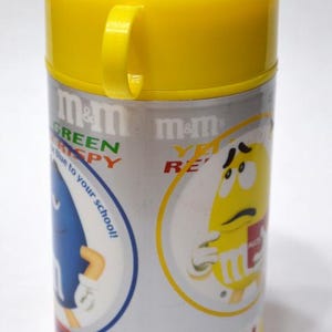 May include: A cylindrical M&M's lunch container with a yellow lid and handle. The container is silver with red accents and features cartoon M&M's characters. Text includes "Green Crispy" and "Yellow Re".