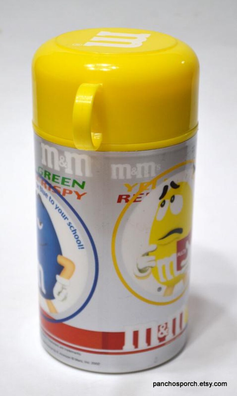May include: A cylindrical M&M's lunch container with a yellow lid and handle. The container is silver with red accents and features cartoon M&M's characters. Text includes "Green Crispy" and "Yellow Re".